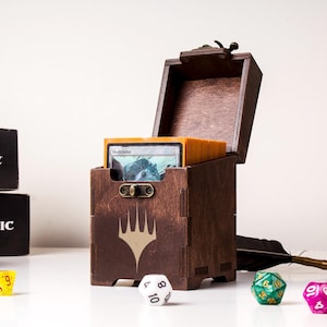May include: A dark brown wooden deck box with a hinged lid and a metal latch, featuring a light-colored Magic: The Gathering logo on the front. The box is filled with a stack of trading cards in orange sleeves, surrounded by various multi-sided dice.