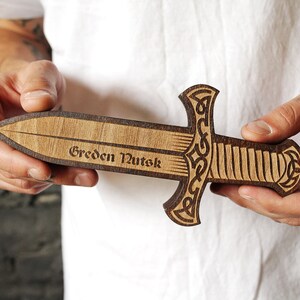 Pode incluir: Uma espada de madeira com um design de n&oacute; celta e a inscri&ccedil;&atilde;o "Greden Nutek".