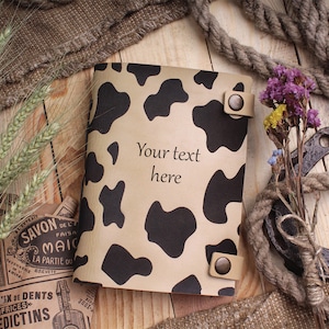 May include: A brown leather notebook with a black and white cow print design. The notebook has two metal snaps and a leather strap. The text "Your text here" is printed on the front cover.