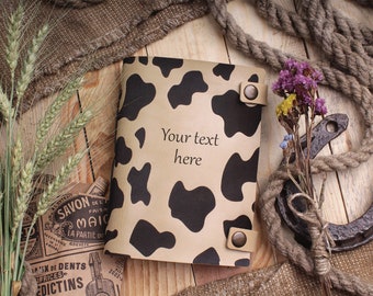 May include: A brown leather notebook with a black and white cow print design. The notebook has two metal snaps and a leather strap. The text "Your text here" is printed on the front cover.
