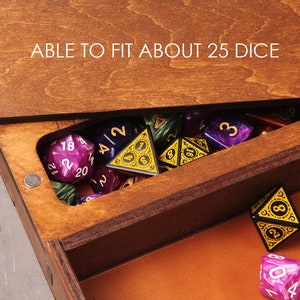 May include: A wooden box with a hinged lid, open to reveal a collection of colorful dice. The lid has the text "ABLE TO FIT ABOUT 25 DICE" engraved on it. The box is filled with dice of various colors, including purple, green, and black.