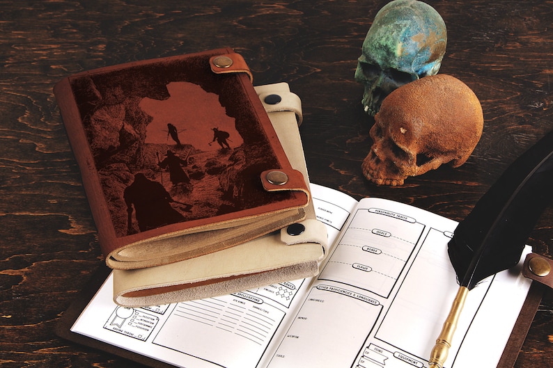Op de afbeelding: Een bruin leren notitieboek met een gegraveerde afbeelding van figuren in een grot. Een ander cr&egrave;mekleurig notitieboek is zichtbaar. Een open notitieboek, een veer en twee decoratieve schedels zijn ook aanwezig.