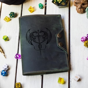 May include: A dark green leather journal featuring a dragon and sword design, secured with two brass press studs. A feather quill, dice, and skull decorations surround the journal on a white wooden surface. The journal is a fantasy-themed item.