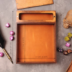 May include: A wooden box with a brown leather interior, measuring 6 2/3 inches by 6 2/3 inches by 9 1/2 inches. The box is open and empty, with a black feather pen resting on the table next to it. There are several dice scattered around the box.