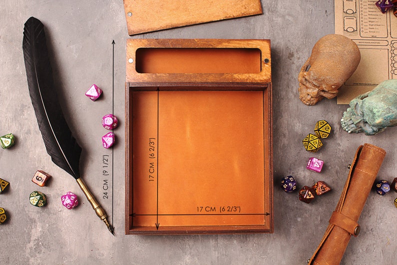 May include: A wooden box with a brown leather interior, measuring 6 2/3 inches by 6 2/3 inches by 9 1/2 inches. The box is open and empty, with a black feather pen resting on the table next to it. There are several dice scattered around the box.