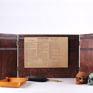 May include: A wooden tabletop game screen with a dark stain, brass hardware, and a paper insert with text. A leather tray, feather quill, and two skulls are in front of the screen.