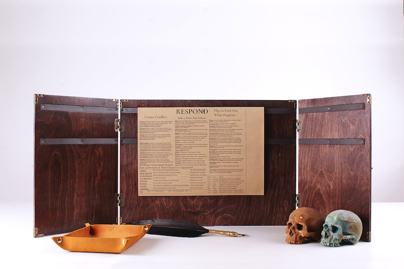 May include: A wooden tabletop game screen with a dark stain, brass hardware, and a paper insert with text. A leather tray, feather quill, and two skulls are in front of the screen.