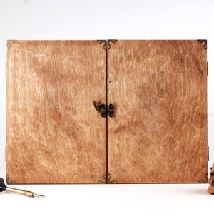 May include: A wooden box with two doors, featuring a dark metal clasp and corner accents. The box has a natural wood grain finish. A skull, a feather quill, and a small box are next to the wooden box.