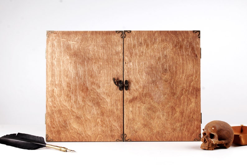 May include: A wooden box with two doors, featuring a dark metal clasp and corner accents. The box has a natural wood grain finish. A skull, a feather quill, and a small box are next to the wooden box.