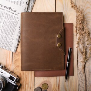 May include: A brown leather bound journal with a flap closure and two brass buttons. The journal is lying on a wooden surface with a pen, a newspaper, a camera, and some coins.