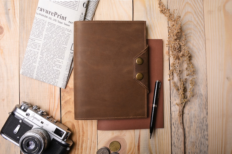 May include: A brown leather bound journal with a flap closure and two brass buttons. The journal is lying on a wooden surface with a pen, a newspaper, a camera, and some coins.