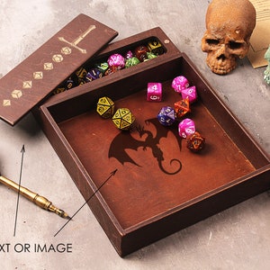 May include: A wooden box with a hinged lid, featuring a sword design on the lid and a dragon silhouette on the inside. The box is filled with colorful dice, and there are two skulls and a leather roll on the table.