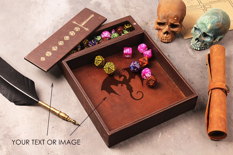 May include: A wooden box with a hinged lid, featuring a sword design on the lid and a dragon silhouette on the inside. The box is filled with colorful dice, and there are two skulls and a leather roll on the table.