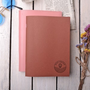 May include: Two leather-bound journals, one pink and one brown, with a hot air balloon logo on the brown journal. The journals are on a white wooden surface with a bouquet of dried flowers.