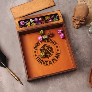 May include: A wooden box with a leather interior and a lid that opens to reveal a compartment for dice. The box has a brown leather interior with the text "Don't Worry I Have A Plan" and a graphic of a 20-sided die. The box is surrounded by dice, a quill pen, and two skull figurines.