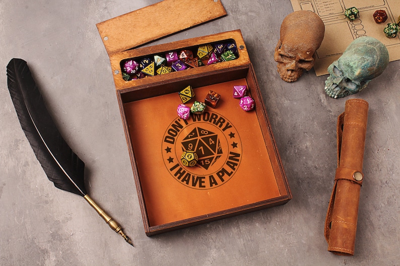 May include: A wooden box with a leather interior and a lid that opens to reveal a compartment for dice. The box has a brown leather interior with the text "Don't Worry I Have A Plan" and a graphic of a 20-sided die. The box is surrounded by dice, a quill pen, and two skull figurines.