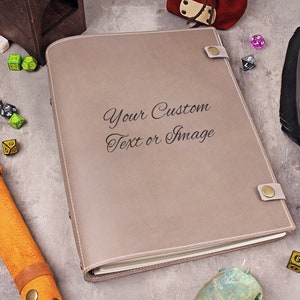 Puede incluir: Un diario encuadernado en cuero gris con detalles de costura blanca y cierre de botón dorado. El diario tiene el texto "Your Custom Text or Image" grabado en la portada.