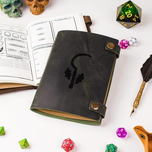 May include: A dark green leather journal with a black sickle and oak leaves graphic, fastened with a strap and button. An open notebook, dice, and a quill pen are also visible, suggesting a role-playing game theme.