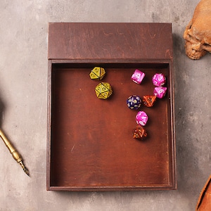 May include: A wooden box with a lid open, revealing a collection of colorful dice. The dice are in various shapes and sizes, including a d20, d12, d10, d8, d6, and d4. The box is sitting on a gray surface.