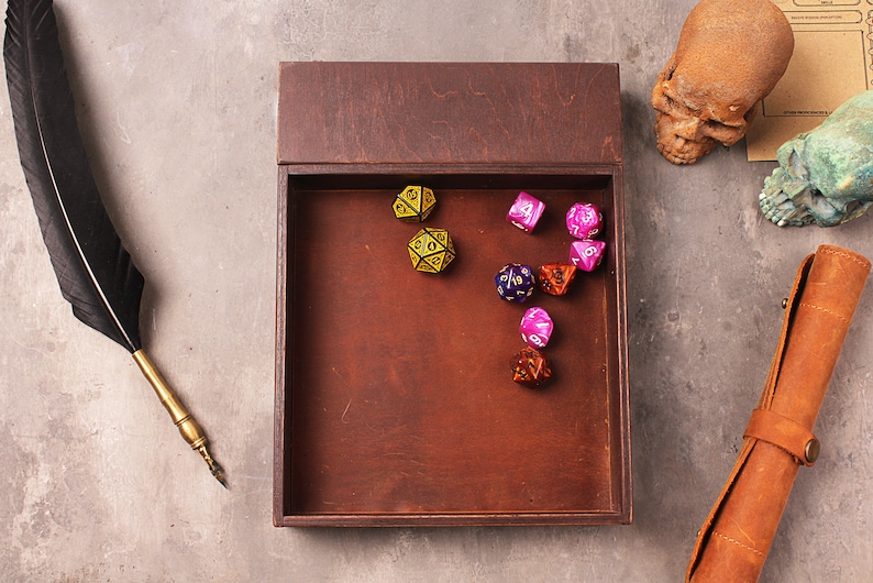 May include: A wooden box with a lid open, revealing a collection of colorful dice. The dice are in various shapes and sizes, including a d20, d12, d10, d8, d6, and d4. The box is sitting on a gray surface.