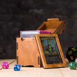 Puede incluir: Una caja de almacenamiento de cartas de madera con tapa con bisagras, que contiene una baraja de cartas. Una carta enmarcada se apoya contra la caja, con varios dados esparcidos. La caja y el marco son de madera marrón claro.
