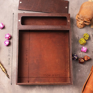 May include: A wooden box with a lid, measuring 17 inches by 17 inches, for storing and rolling dice. The box is made of brown wood and has a brown leather interior. The box is surrounded by dice and a quill pen.