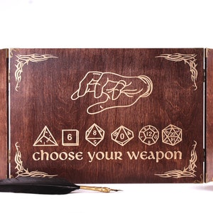 Może przedstawiać: Drewniana skrzynia z zawiasowym wiekiem, przedstawiająca wyciągniętą rękę i napis "choose your weapon". Skrzynia ma brązowe wykończenie z drewna i jest ozdobiona złotymi kostkami do gry i stylizowanym wzorem.