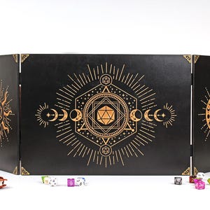 Może przedstawiać: Czarny, trzypanelowy ekran stołowy ze złotymi wzorami niebieskimi, w tym słońcami, księżycami i kształtami geometrycznymi. Ekran jest przeznaczony do gier fabularnych na stole. Kości i skórzany notatnik na pierwszym planie.