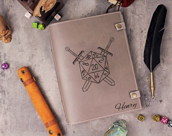 May include: A gray leather journal with a black and white illustration of a twenty-sided die with two swords crossed behind it. The journal has a flap closure with a metal button and the name "Henry" is embossed on the front.