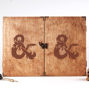 May include: Wooden box with a dragon and ampersand design. The box has a hinged lid with a clasp. The design is etched in brown on the wooden surface. The box is likely for storing items related to fantasy role-playing games.