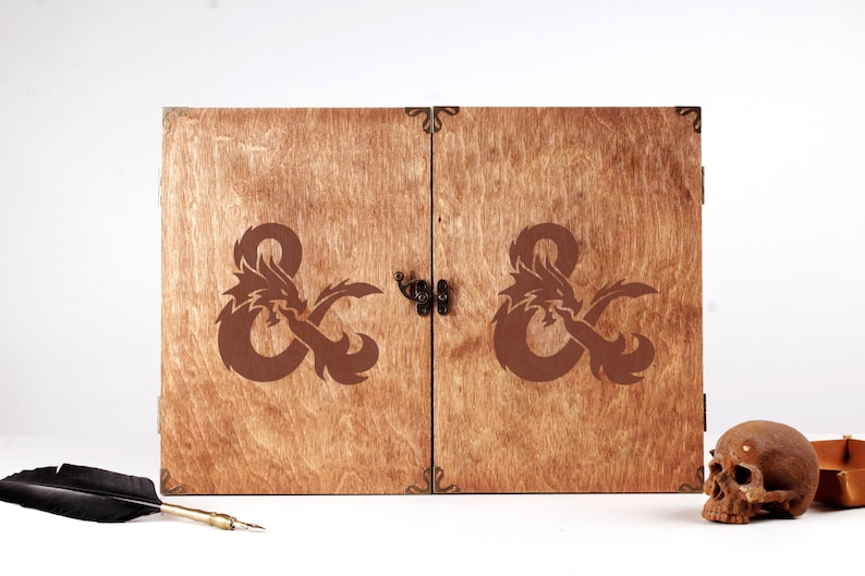 May include: Wooden box with a dragon and ampersand design. The box has a hinged lid with a clasp. The design is etched in brown on the wooden surface. The box is likely for storing items related to fantasy role-playing games.
