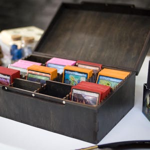 May include: A dark brown wooden storage box with a hinged lid, containing multiple rows of organized trading cards in various colored sleeves. Next to the box are two black Magic The Gathering branded card boxes with white text.