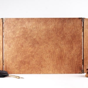 May include: A wooden, three-panel folding screen with brass hinges and clasps. A black feather quill and a small skull are placed in front of the screen. The wood has a warm, natural tone.