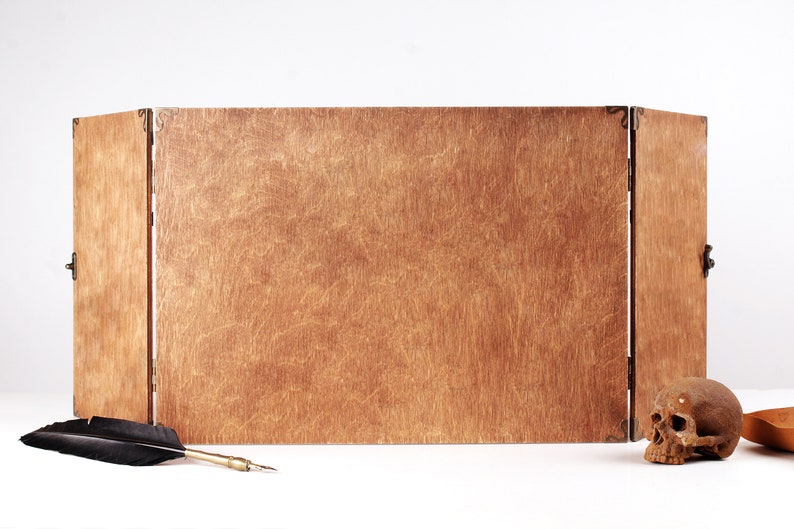 May include: A wooden, three-panel folding screen with brass hinges and clasps. A black feather quill and a small skull are placed in front of the screen. The wood has a warm, natural tone.
