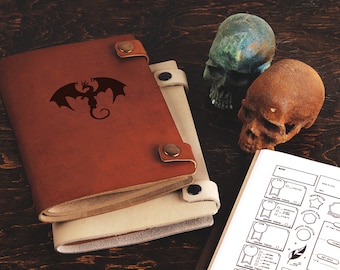 May include: A brown leather-bound journal with a dragon silhouette on the cover. The journal is closed and has a white interior. Two decorative skulls are on a wooden surface next to the journal.