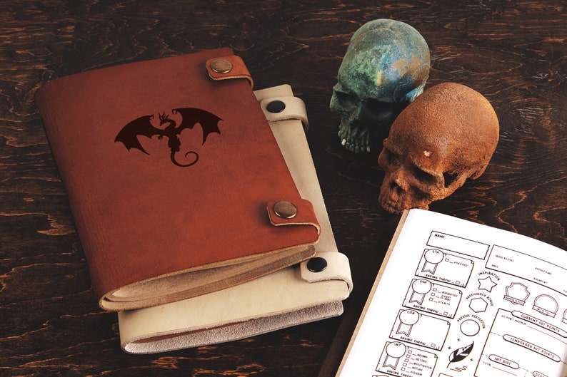 Op de afbeelding: Twee leren gebonden tijdschriften, &eacute;&eacute;n bruin en &eacute;&eacute;n beige, met een draken silhouet. Twee decoratieve schedels, &eacute;&eacute;n groen en &eacute;&eacute;n bruin, zijn in de buurt geplaatst. Een open boek met personagebladen is ook zichtbaar.