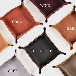 Könnte beinhalten: Eine Auswahl an Leder-Ablageschalen in verschiedenen Farben, darunter Beige, Cognac, Ziegel, Schokolade, Grau, Olive und Wein. Jede quadratische Schale hat abgerundete Ecken und genähte Kanten. Die Farbbezeichnungen sind auf jeder Schale zu sehen.