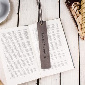 May include: A gray leather bookmark with the text "Fuck off I'm reading" is placed in an open book. A cup of coffee and other books are on a white wooden surface. Dried flowers and a newspaper are also visible.