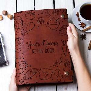 Peut inclure: Un livre de recettes en cuir marron avec un dessin à la main d'aliments et le texte "Your Name RECIPE BOOK" sur la couverture.