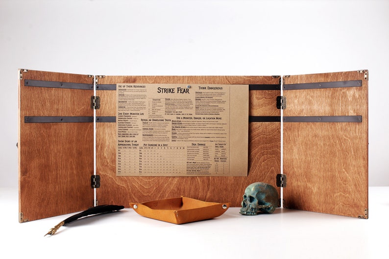 May include: A wooden tabletop gaming screen with a brown leather tray, a teal skull, and a feather quill. The screen has a printed chart that reads "Strike Fear". The screen is made of wood and has metal accents.