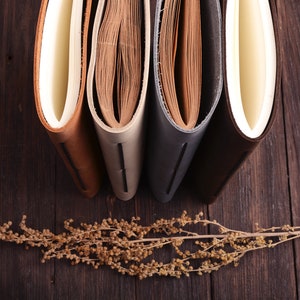 K&ouml;nnte beinhalten: Vier ledergebundene Journale mit verschiedenen Farbt&ouml;nen, darunter Braun, Beige, Grau und Braun, sind nebeneinander auf einer Holzoberfl&auml;che angeordnet. Ein getrockneter Blumenzweig liegt vor den Journalen.