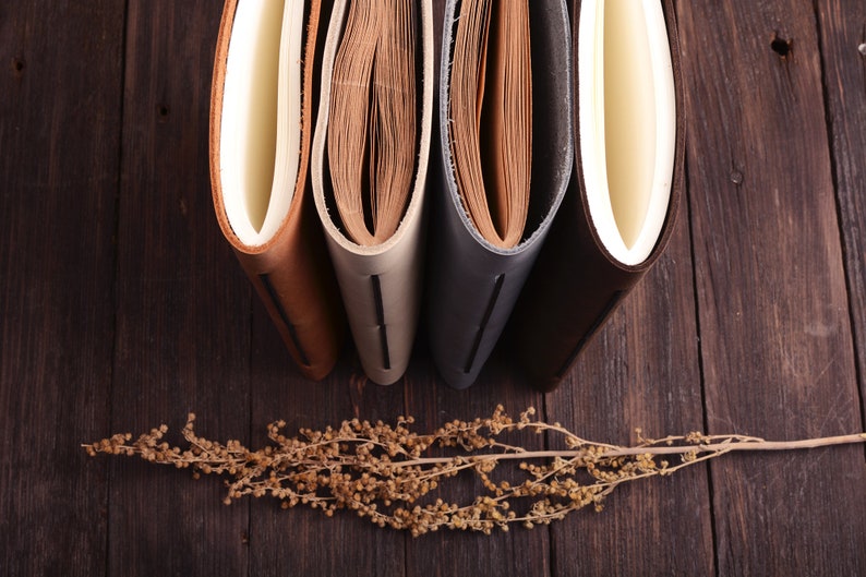 K&ouml;nnte beinhalten: Vier ledergebundene Journale mit verschiedenen Farbt&ouml;nen, darunter Braun, Beige, Grau und Braun, sind nebeneinander auf einer Holzoberfl&auml;che angeordnet. Ein getrockneter Blumenzweig liegt vor den Journalen.