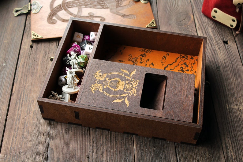 May include: A wooden box with a hinged lid, containing dice, miniature figures, and a map. The box has a decorative design on the front and a compartment for storing dice.