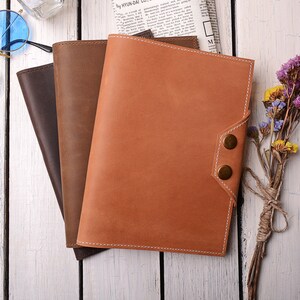 May include: Three leather-bound journals in brown, dark brown, and orange, stacked on a white wooden surface. The journals have a stitched edge and a flap closure with brass buttons.