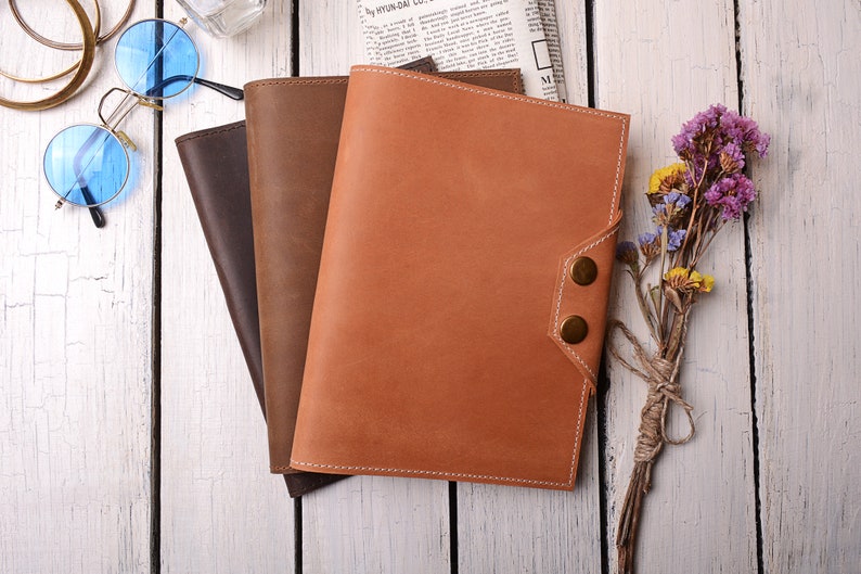 May include: Three leather-bound journals in brown, dark brown, and orange, stacked on a white wooden surface. The journals have a stitched edge and a flap closure with brass buttons.