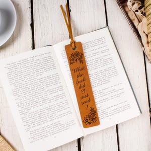 May include: A brown leather bookmark with the text "What the fuck did I just read?" and floral designs. The bookmark is in an open book, with a cup of coffee and other books nearby. The bookmark has a leather cord.
