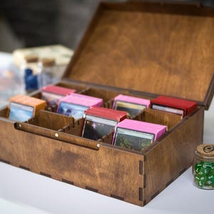 May include: A wooden card storage box with multiple compartments, holding various card decks with pink and red edges. The box is open, revealing the cards inside. Small glass jars with dice are in the foreground.