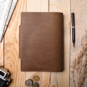 May include: A brown leather notebook cover with a flap on the left side. The cover is on a wooden surface with a pen, coins, and a camera.