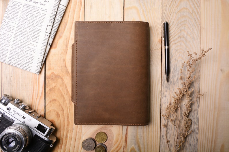 May include: A brown leather notebook cover with a flap on the left side. The cover is on a wooden surface with a pen, coins, and a camera.