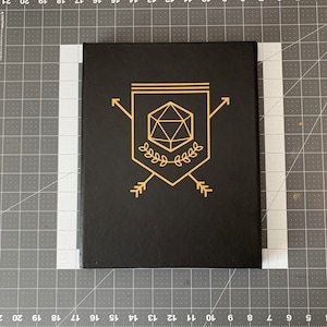 May include: A black notebook with a gold design of a 20-sided die, arrows, and leaves. The design is centered on the cover.
